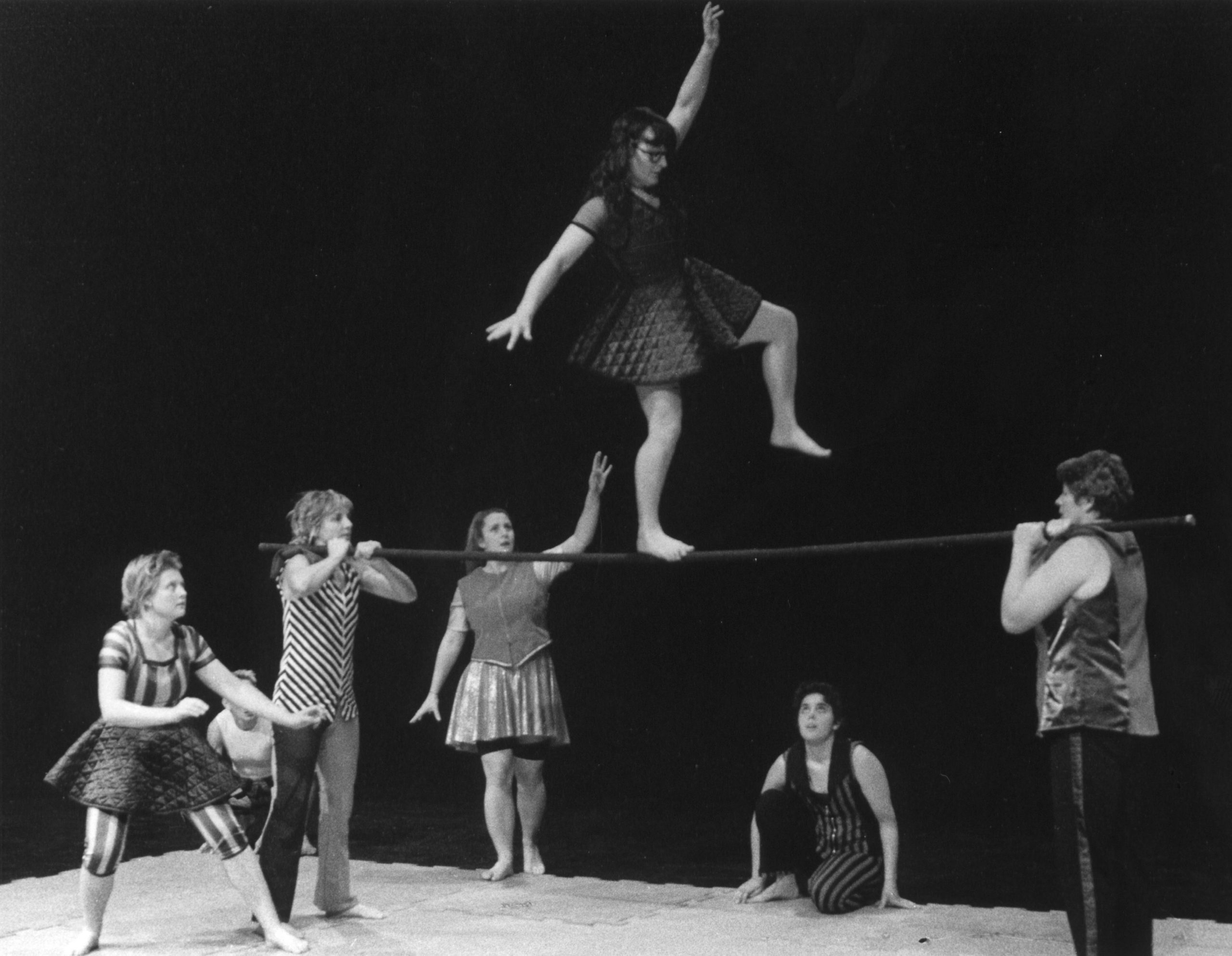 A group of people standing by as one of the women walks along a pole.