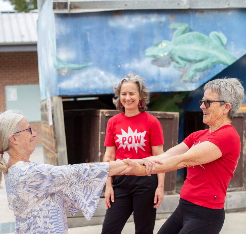 two older people balance in a counter balance while another wearing a POW t shirt smiles and watches